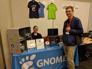 A photo of the GNOME booth at LFNW. Behind the booth sits one person, and another is standing in front of it. The table is blue.