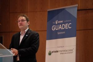 A photo of Neil McGovern, Ecexutive Director of the GNOME Foundation in August 2019. He is wearing a suit. Behind him is a sign that says "GUADEC" and "Private Internet Access."