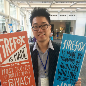 A photo of Sammy Fung holding up two firefox signs. He is wearing a suit jacket and a blue collared shirt. He has glasses and his hair is sticking up.