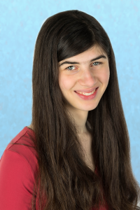 A young woman, against a blue background, wearing a red shirt. She has long, dark brown hair.