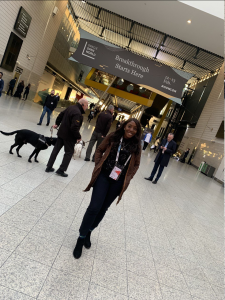 A photo of Regina, who is smartly dressed in black with a green jacket, standing in a conference center. Behind her is a man with a dog.