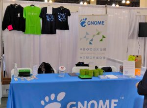 A photo of the GNOME booth, featuring a blue GNOME table cloth and exciting GNOME swag, including bugs, t-shirts, and a tote bag.