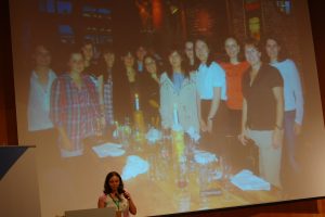 A photo of Marina Zhurakhinskaya in front of a photo of a group of women from the GNOME Women's Dinner.