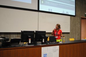 A woman stands behind a desk, looking up at slides. She has a microphone in her hand and is giving a presentation.