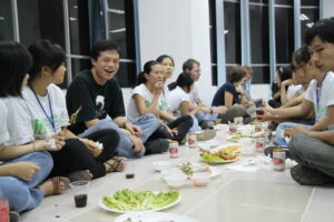 A photo of a group of people in matching shirts, sitting on a table making spring rolls.