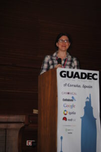 A person in a black, pink, and white flannel behind a speaker podium with a sign that says "GUADEC."