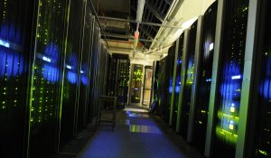 A dark server room, filled with computer stacks emitting eerie blue and green light.