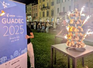 A birthday cake with sparklers stands next to a sign saying GUADEC 2025
