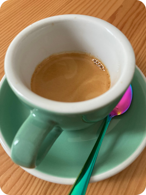 Light green demitasse with saucer and a spoon shining in rainbow colors. The cup contains espresso with brown crema.